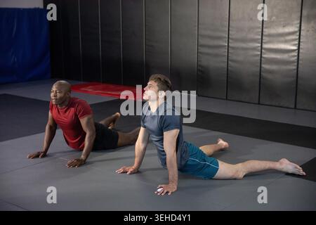 Deux hommes allongés sur des tapis emboîtants, faisant l'étirement du dos couché dans la salle de gym d'arts martiaux portant des shorts. Athlètes, partenaires, entraînement, fitness, concentration, travail d'équipe, flexibilité Banque D'Images
