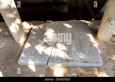 Vieste - Pouilles - Italie - 29-08-2025 : dalle de marbre fissuré avec inscription latine sculptée repose sur un sol en pierre entre les colonnes, avec des ombres de feuilles et la lumière du soleil à travers elle. Banque D'Images