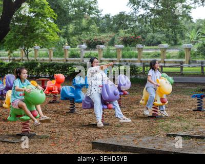 Heureuse mère asiatique et ses deux filles chevauchant des cavaliers de printemps colorés à l'aire de jeux extérieure. Banque D'Images
