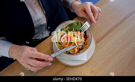 Femme d'affaires senior mangeant de la salade dans le café, légumes vibrants avec fourchette et couteau sur la table en bois, bol en porcelaine, filtre naturel de la lumière du soleil Banque D'Images
