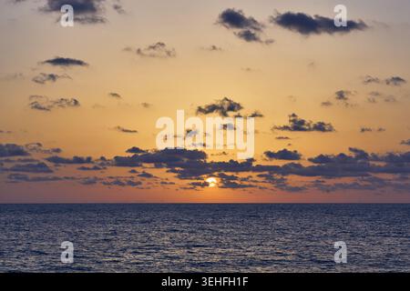 Soleil plongeant sous l'horizon, peignant le ciel avec des couleurs chaudes et des nuages dispersés sur l'océan calme Banque D'Images