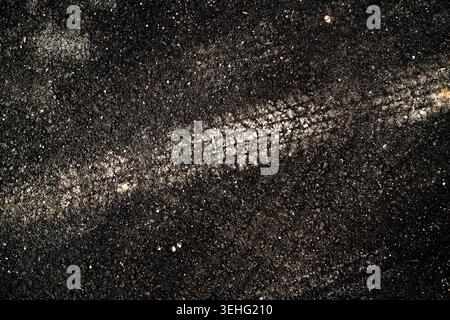 Gros plan d'une voie à pneu unique enfoncée dans une chaussée asphaltée sombre, mettant en évidence le motif de la bande de roulement, la texture granuleuse et le contraste. Vue de dessus. Banque D'Images