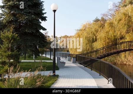 Un parc serein avec un sentier sinueux, un pont en bois et des arbres luxuriants avec feuillage d'automne. Un lampadaire se tient le long du chemin, améliorant le tranqui Banque D'Images