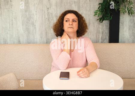 Femme d'âge moyen assise à une table regardant réfléchi et attendant avec le téléphone devant elle Banque D'Images