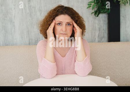 Femme tenant ses tempes avec mal de tête et stress tout en étant assise à une table dans un salon Banque D'Images