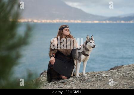 Une femme est assise à côté d'un chien Husky sibérien sur les rochers côtiers. Le paysage de montagne de mer bleue et la posture détendue créent une atmosphère paisible de style de vie de voyage Banque D'Images
