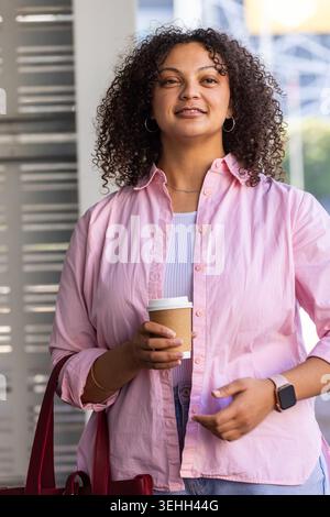 Femme afro-américaine debout dans le hall tenant une tasse de café en papier, portant une chemise rose et un fourre-tout rouge Banque D'Images