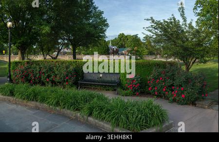 Un banc de parc en métal noir se trouve devant une haie verte avec des roses rouges en fleurs près d'un pont de pierre et d'une passerelle pavée à Baker Park, Frederick, Maryla Banque D'Images