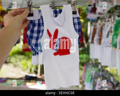 Une personne tenant un cintre affichant un maillot blanc pour enfants fait à la main avec un patch de lapin rouge et un short Vichy bleu assorti. Banque D'Images