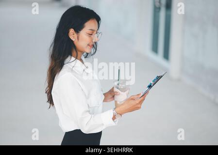 Vue de côté d'une femme professionnelle souriante avec du café glacé et une planche à pince, soulignant la routine de bureau, pause de travail décontractée, humeur positive, Li d'affaires Banque D'Images