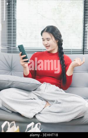 Jeune femme asiatique souriant au smartphone sur le canapé à la maison avec des sacs à provisions au premier plan, vérifiant l'application d'achat en ligne, la mise à jour de la commande de commerce électronique et creuser Banque D'Images