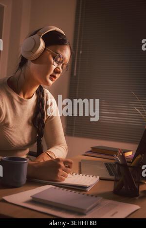 Jeune femme asiatique portant des écouteurs et des lunettes tout en écrivant des notes par ordinateur portable dans le bureau à domicile la nuit, représentant l'apprentissage en ligne, le travail à distance, con Banque D'Images