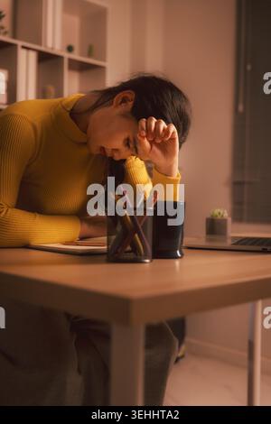 Jeune femme asiatique épuisée couvrant les yeux tout en étant assise par ordinateur portable dans un bureau à domicile confortable la nuit, illustrant l'épuisement professionnel, le stress d'étude, la fatigue au travail, mort Banque D'Images