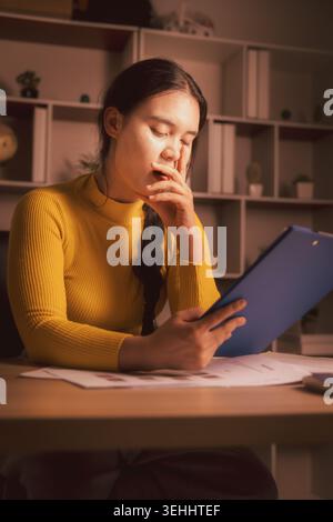 Jeune femme asiatique endormie bâillant tout en examinant des documents de presse-papiers dans un bureau à domicile confortable la nuit, illustrant la fatigue de la paperasse, le stress des heures supplémentaires, de Banque D'Images