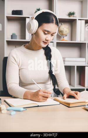 Jeune femme asiatique étudiant à la maison avec des écouteurs, lisant un livre, prenant des notes et utilisant un ordinateur portable au bureau. Apprentissage en ligne, éducation ciblée, à distance Banque D'Images