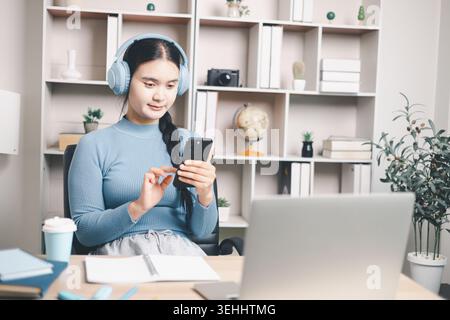 Jeune femme asiatique portant des écouteurs et utilisant un smartphone à côté d'un ordinateur portable au bureau à la maison. Apprentissage mobile, cours en ligne, étude à distance, enseignement numérique a Banque D'Images