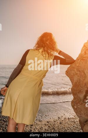 Femme coucher de soleil femme de plage en robe jaune appuyée sur un rocher regardant les vagues de l'océan pendant l'heure d'or avec l'espace de copie Banque D'Images