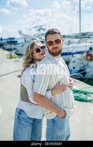 Un couple se tient à une marina, entouré de divers bateaux et de l'eau. La femme enroule ses bras autour de l'homme alors qu'il regarde vers la caméra. Ils ont tous les deux w Banque D'Images