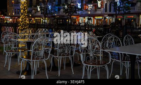 Chaises de café vides en fer forgé blanc et tables à côté d'un canal illuminé festivement avec des bâtiments de style européen la nuit à Grand World, Phu Quoc, Vietnam. Banque D'Images