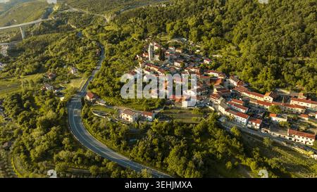 Vue aérienne du village historique de Crni Kal avec ses maisons au toit rouge et sa tour d'église nichée parmi les collines verdoyantes et le viaduc de Crni Kal Banque D'Images