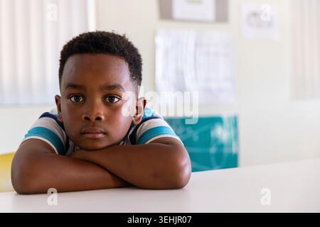 Garçon afro-américain assis à un bureau blanc, se reposant les bras pliés et regardant la caméra dans la classe. Étudiant, calme, concentration, éducation, sarcelle, jaune, bulletin Banque D'Images