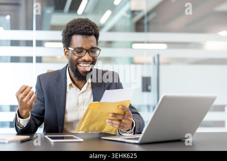 Jeune homme professionnel lisant des nouvelles triomphales à partir d'une enveloppe, se sentant excité et célébrant un résultat réussi ou une promotion dans un espace de travail moderne Banque D'Images