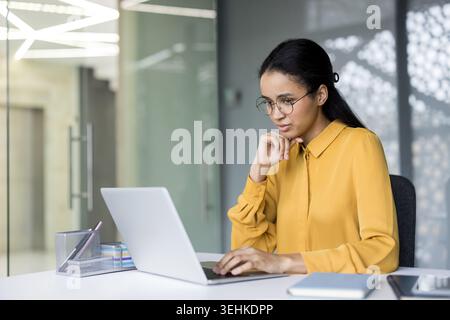 Jeune femme d'affaires portant des lunettes et une chemise jaune se concentrant sur son travail, tapant sur un ordinateur portable à un bureau moderne, incarnant le dévouement et l'efficacité Banque D'Images