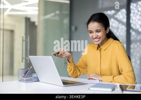 Femme professionnelle souriante faisant des gestes pendant une visioconférence sur son ordinateur portable, collaborant à distance depuis un espace de travail moderne, confiante et engagée dans une réunion virtuelle Banque D'Images