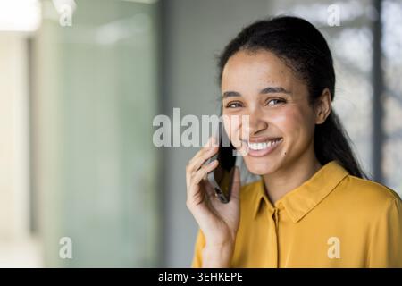 Jeune femme d'affaires debout dans un bureau, tenant un smartphone à son oreille, souriant brillamment tout en s'engageant dans une conversation agréable, symbolisant la communication et la connexion d'entreprise modernes Banque D'Images