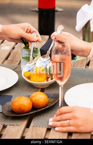 Un gros plan de deux personnes utilisant des cuillères pour partager un bol de sorbet à la mangue sur une table en bois extérieure, avec des cocktails et des petites pâtisseries dans le Forward Banque D'Images