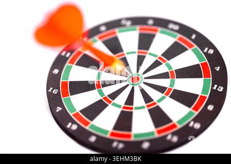 Fléchette avec un vol orange est coincée dans le logo de la cible d'un jeu de fléchettes, qui a des segments noirs, blancs, rouges et verts, définir sur un fond blanc Banque D'Images