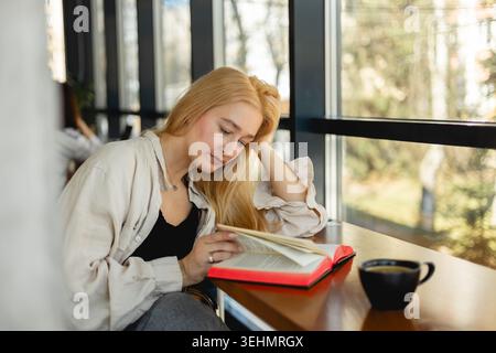 Une femme aux cheveux blonds s'assoit confortablement dans un café près d'une fenêtre, engrossée dans la lecture d'un livre. Une tasse de café vapeur repose sur la table, créant un Banque D'Images