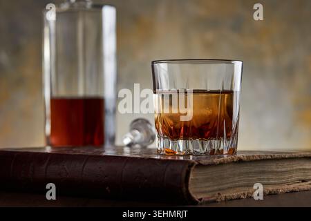 Gros plan de bourbon de whisky sur les rochers dans un gobelet coupé, sur un livre de cuir usé avec une bouteille floue en arrière-plan et une lumière de bar chaude et d'humeur changeante Banque D'Images