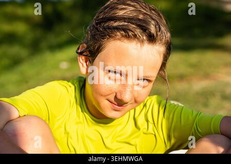 Portrait en gros plan d'un garçon aux cheveux mouillés souriant dans une chemise lumineuse, se relaxant sur l'herbe au bord d'un lac par une journée d'été ensoleillée, profitant des loisirs en plein air et des vacances Banque D'Images