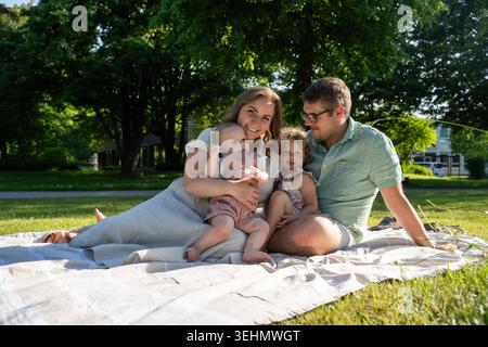 Les parents souriants se détendent sur une couverture de pique-nique dans un parc ensoleillé, tenant une petite fille et câlinant une fille en bas âge, capturant l'amour, le bonheur et l'été Banque D'Images