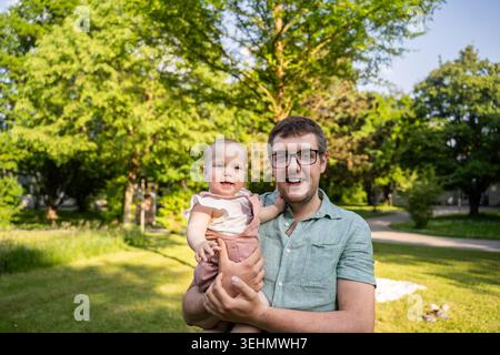 Le père souriant tient sa petite fille à l'extérieur dans un parc verdoyant par une journée d'été ensoleillée, capturant l'amour, le bonheur et le temps chaud en famille ensemble dans la nature. Banque D'Images