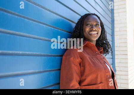 Portrait d'une femme africaine souriante avec des cheveux tressés appuyés contre un obturateur en métal bleu à l'extérieur, portant une veste de rouille dans une lumière naturelle éclatante. Banque D'Images