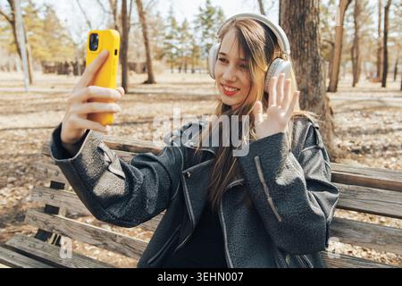 Fille souriante dans une veste chaude prend un selfie avec un smartphone jaune tout en portant des écouteurs circum-aural, assis sur un banc en bois dans un parc ensoleillé Banque D'Images