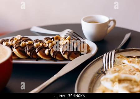 Table de petit déjeuner avec mini crêpes arrosées de chocolat et garnies de noisettes, ainsi que des crêpes à la pomme et au fromage à la crème, servies avec du café frais Banque D'Images