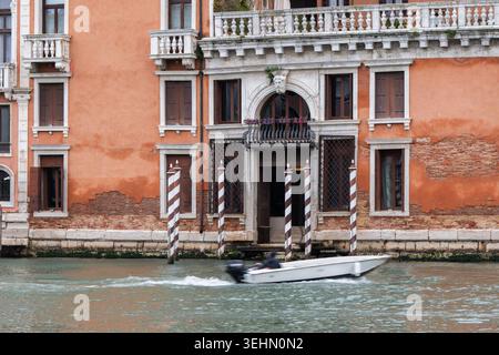 Façade de bâtiment vénitien historique le long d'un canal, avec des fenêtres à volets, balcon en pierre, poteaux d'amarrage rayés, et un petit bateau à moteur passant sur la wa Banque D'Images