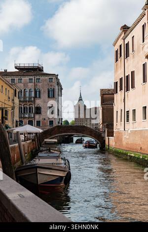 Pittoresque canal de Venise avec bateau amarré et petit bateau à moteur passant sous un pont en arc de brique, encadré par des bâtiments historiques pastels et un ciel bleu doux dedans Banque D'Images