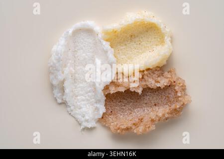 Gros plan de trois frottis de gommage corporel montrant une texture exfoliante crémeuse et granuleuse en blanc, jaune pâle et brun, disposés sur un fond beige doux Banque D'Images