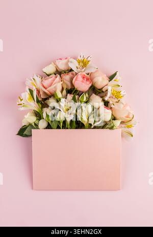 Bouquet pastel doux de roses roses roses et alstroemeria blanche disposés derrière une carte de voeux vierge sur fond rose. Banque D'Images