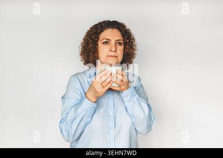 Femme d'âge moyen tenant une tasse à café, portrait réfléchi en chemise bleue sur fond blanc Banque D'Images