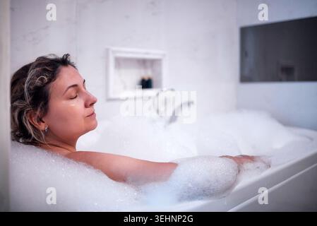 Femme se détend dans un bain moussant à l'intérieur d'une salle de bains moderne, avec carrelage blanc et une baignoire élégante avec un éclairage doux créant un environnement apaisant. Banque D'Images
