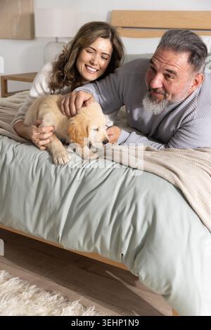 Couple avec un homme mature et une femme adulte moyenne couchée sur le lit, caressant le chiot Golden retriever Banque D'Images