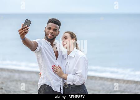 Un couple se tient proche à Playa del Zapillo, Almeria, Espagne, souriant en prenant un selfie. Leurs expressions joyeuses reflètent la plage ensoleillée de Banque D'Images
