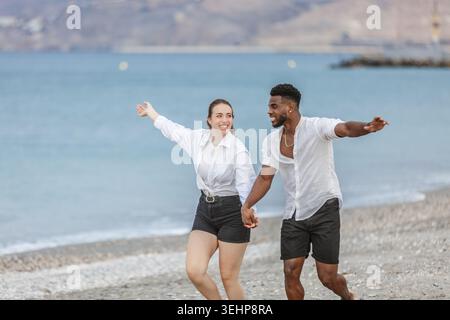Un couple court de manière ludique le long du rivage à Playa del Zapillo, Almeria, Espagne. La femme, vêtue d'une chemise blanche et d'un short, rit joyeusement, whi Banque D'Images