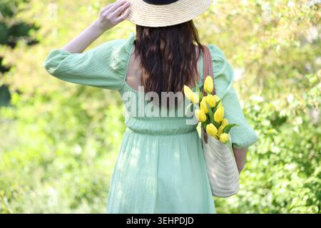 Femme en robe et chapeau de paille tenant un sac en osier avec de belles fleurs de tulipe à l'extérieur le jour ensoleillé, vue arrière Banque D'Images