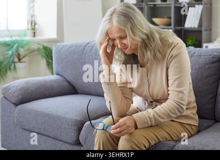 Femme âgée triste avec mal de tête tenant le front et les lunettes dans les mains, assise sur le canapé à la maison Banque D'Images